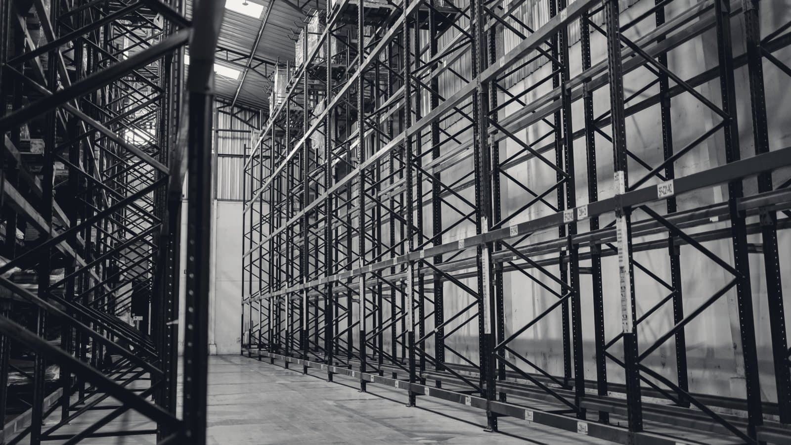 A warehouse aisle lined with tall, mostly empty steel pallet racks, daylight falling from roof lights onto the bare concrete floor.