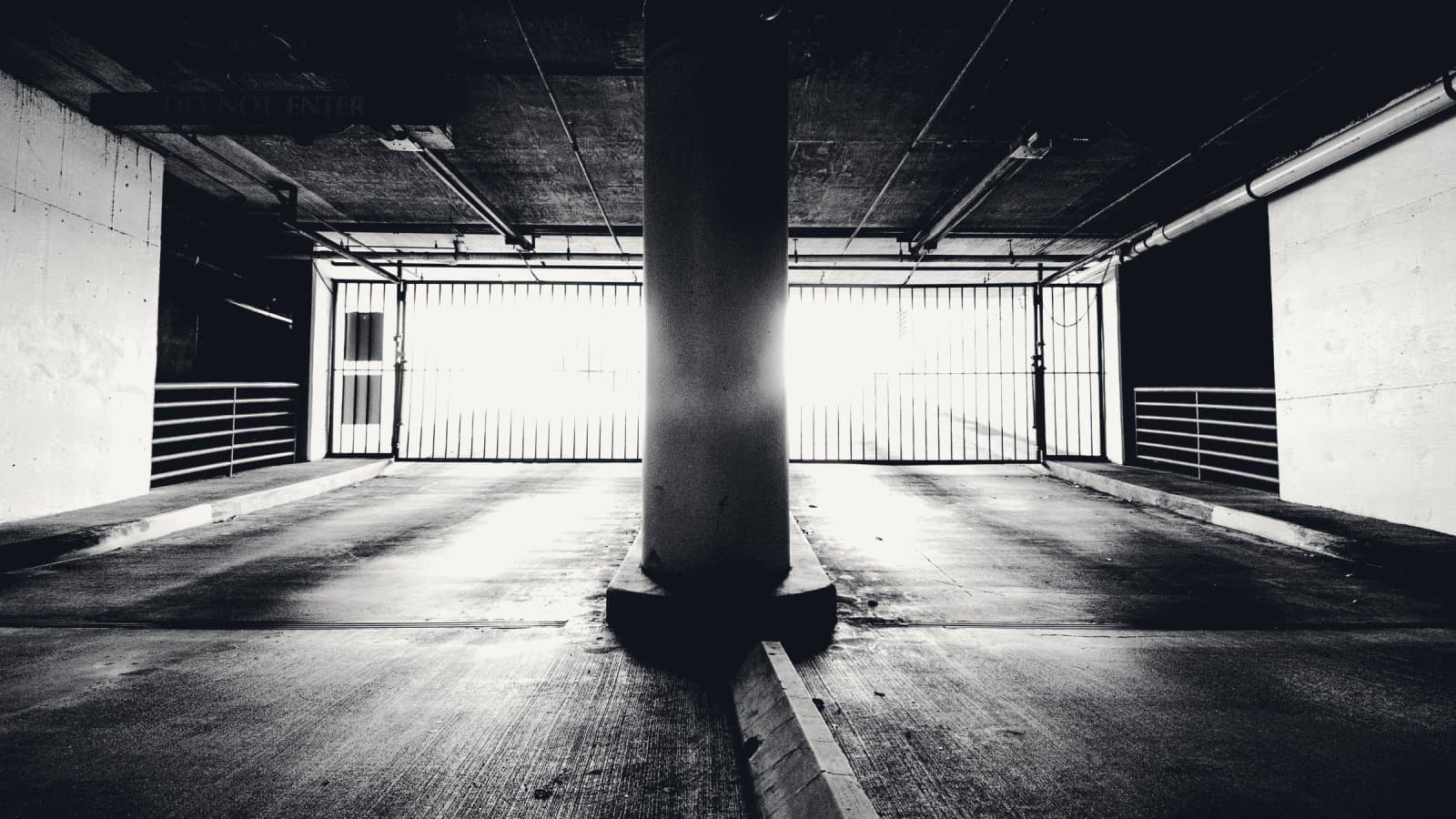 A parking structure seen from the ramp, a low concrete slab and one round column in the foreground, with a barred gate at the exit and bright daylight beyond.
