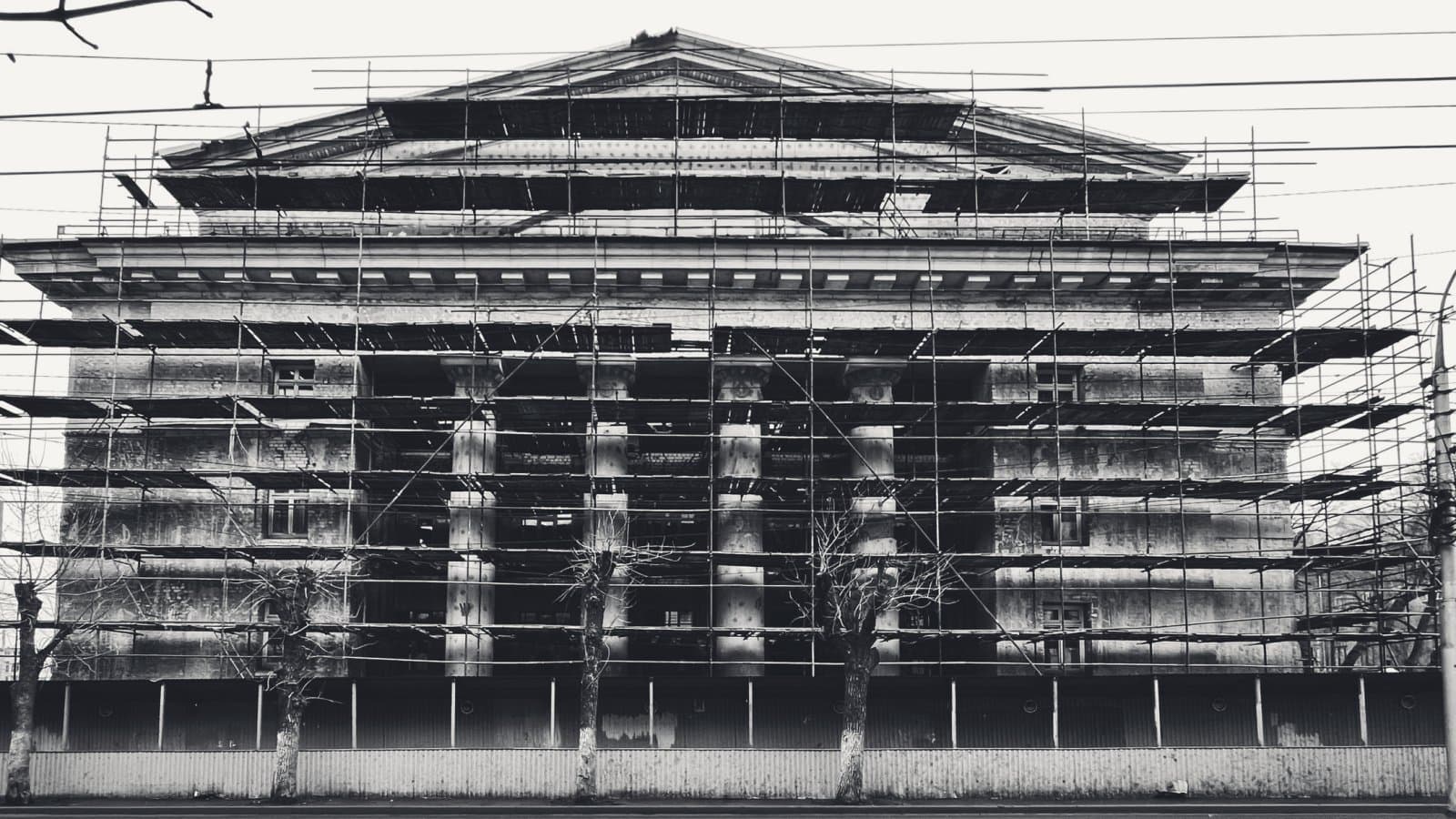 A neoclassical building with a columned portico and pediment wrapped from street level to roofline in scaffolding, bare trees and a corrugated hoarding in front.