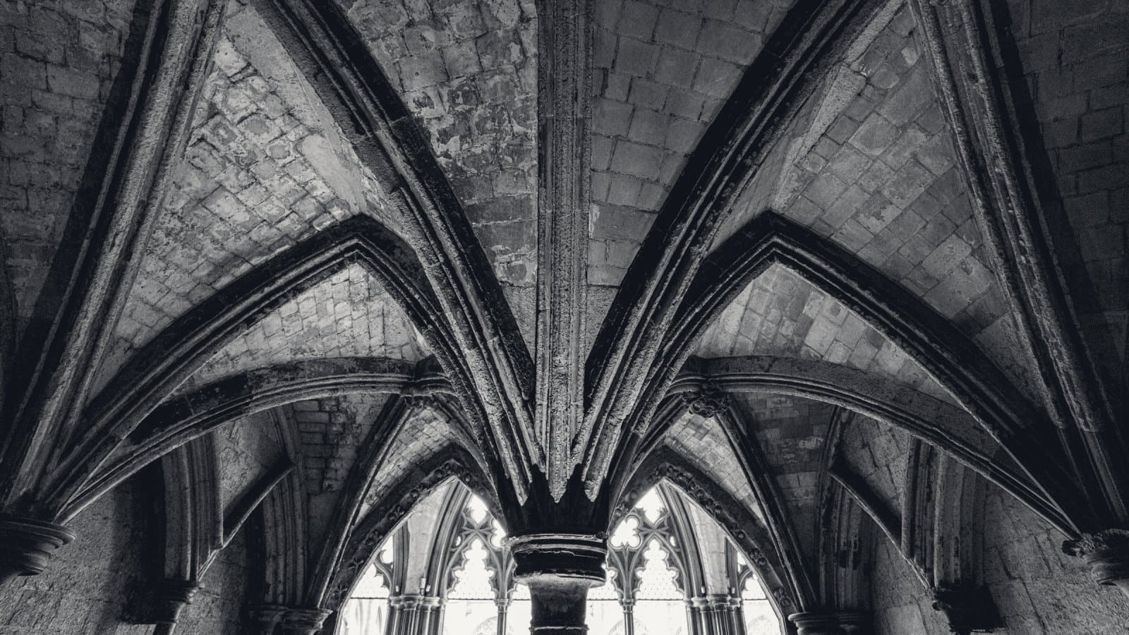 A weathered stone rib vault seen from below, its ribs fanning out from a single column, with daylight through tracery windows behind.