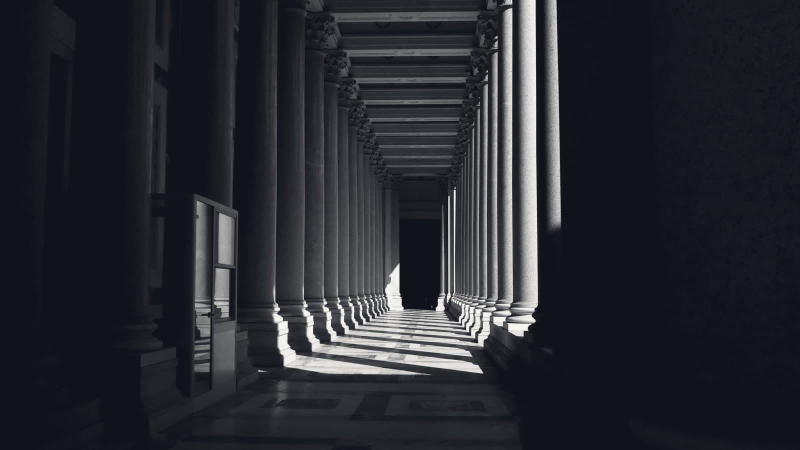 A long colonnade of stone columns receding into shadow, with hard directional light falling across the floor between them.